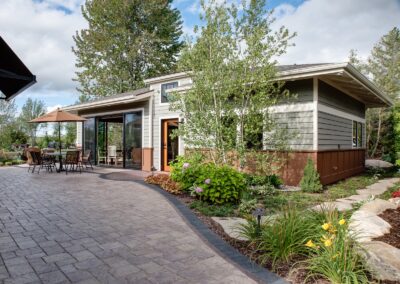 A modern, single-story house with large glass sliding doors and a patio. The patio features a dining set with a large umbrella, surrounded by a lush garden with various plants and flowers. The backdrop includes trees and a clear, partly cloudy sky.